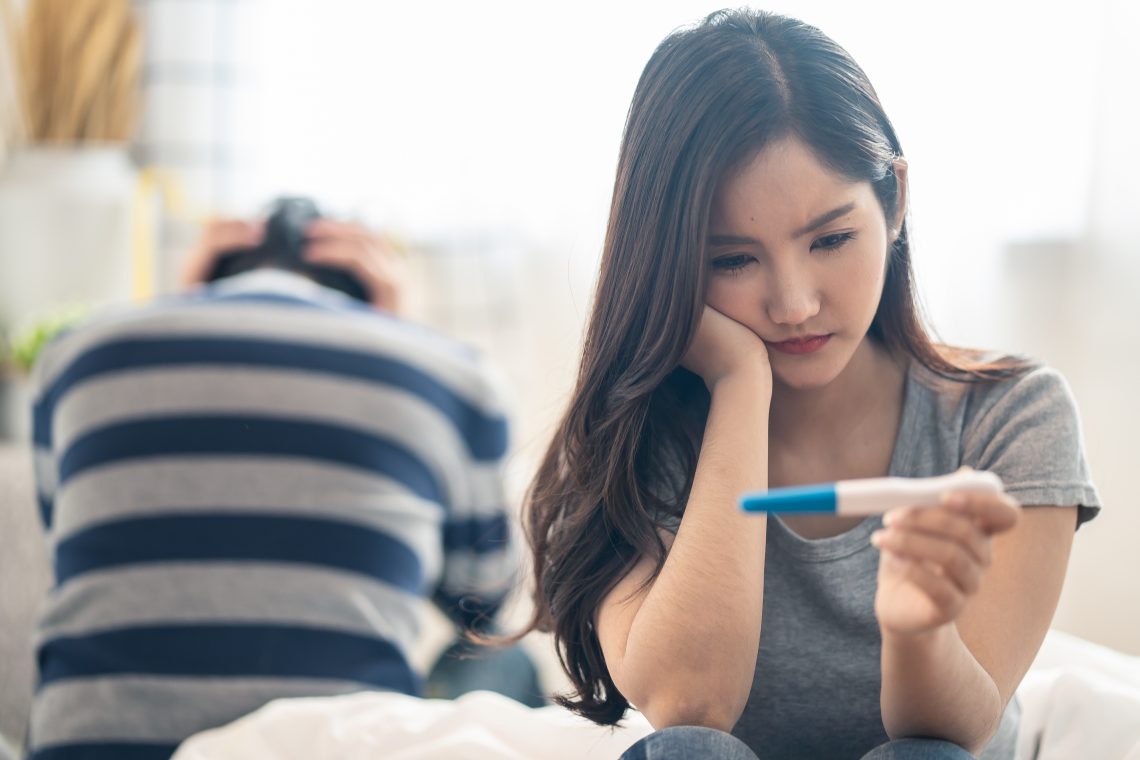 Disappointed Asian woman sitting bed when see the pregnancy test result. Girl feels worry and not ready to be a mother. Man feeling stress about the future kid holding head. Unprepared sexual problem.